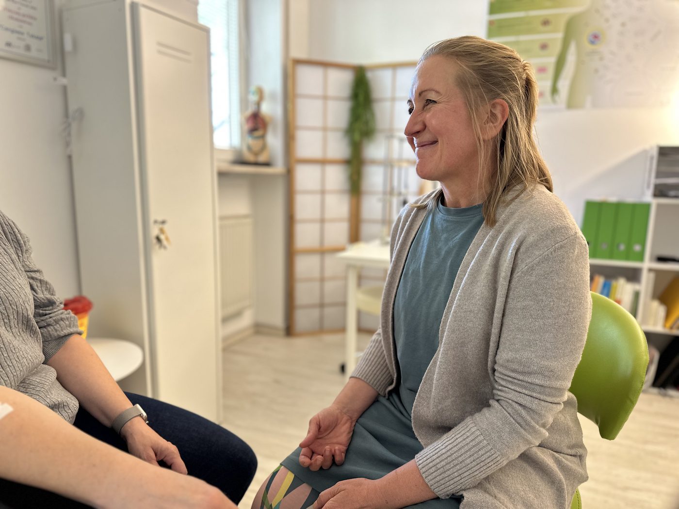 Delia Gröninger sitzt lächelnd im Gespräch mit einer Patientin, von der nur Arme und Knie zu sehen sind.