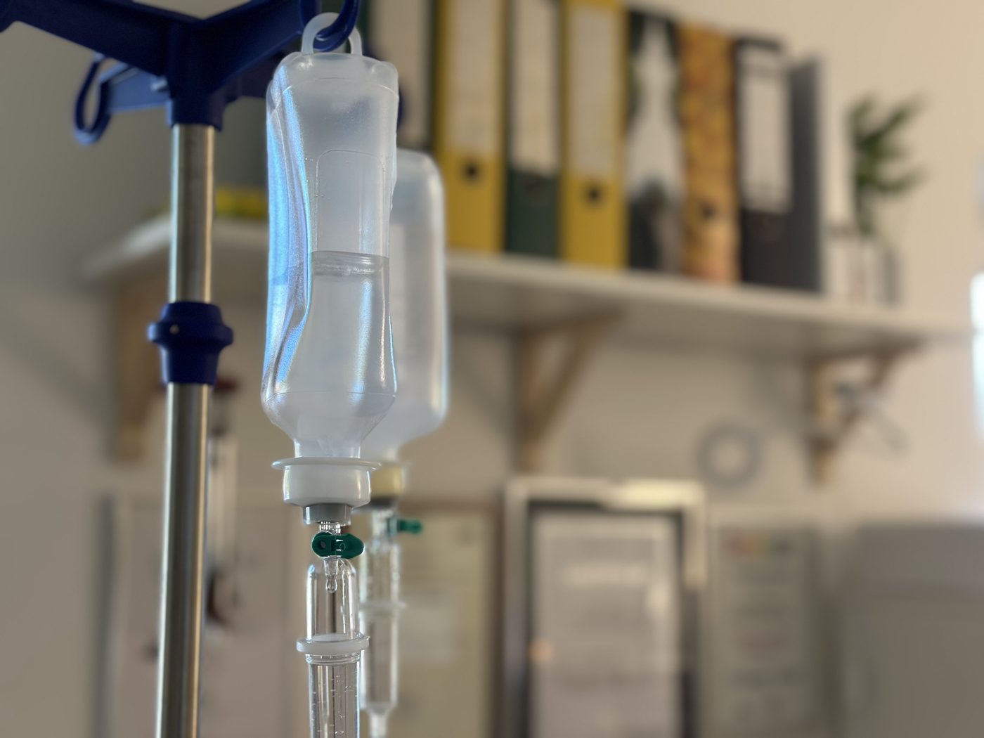 Zwei Infusionsflaschen mit klarer Lösung hängen am Infusionsständer, dahinter unscharf ein Regal mit Ordnern.